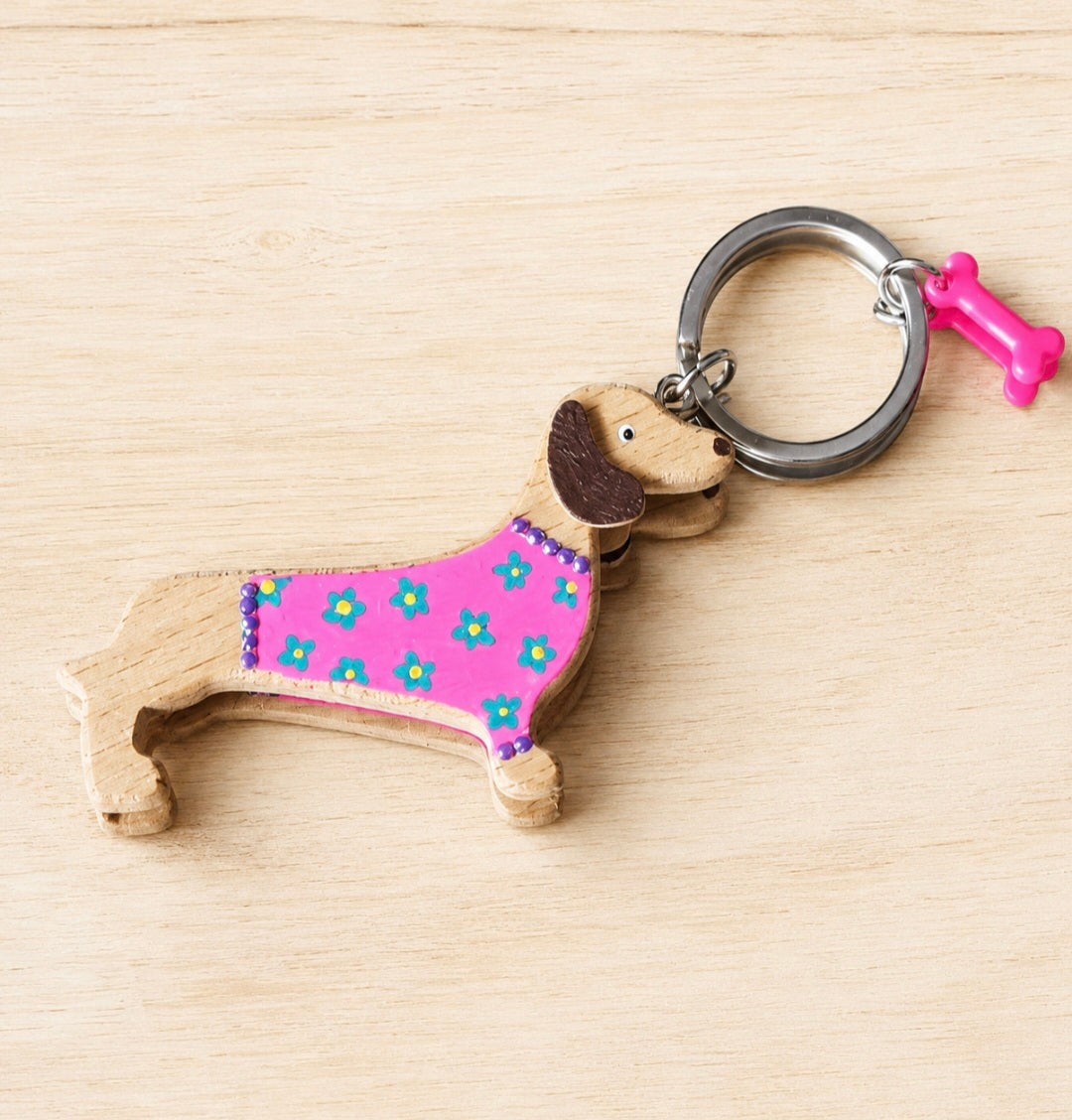 A wooden dachshund-shaped keychain with a pink floral coat, brown ears, and a silver key ring, shown on a light wood background.